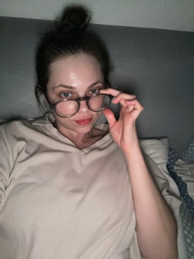 One adult female subject is depicted in a dimly lit indoor setting in Monsummano Terme, Italy. Her dark hair is styled in a high bun. She is wearing a light beige hooded top and round, dark-framed eyeglasses. Her left hand is raised, holding the left temple of the eyeglasses, slightly lowering them from her eyes. Her light-colored eyes are directed at the viewer, and a subtle smirk is visible on her lips, which have a reddish tint. The background consists of a plain dark grey surface, likely a wall or headboard, with a light-colored pillow visible to the subject's right and blue and white patterned bedding visible to her left.