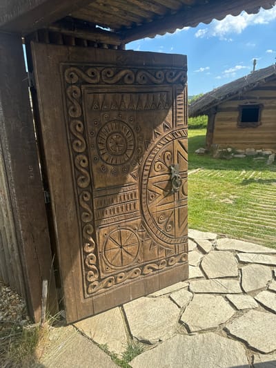 A close-up, eye-level, outdoor shot captures an intricately carved wooden door, weathered and aged, set against a backdrop of a grassy field and a thatched-roof building. The door, adorned with geometric patterns and circular motifs, is partially open, revealing a glimpse of a rustic wooden structure. Sunlight casts shadows across the door, highlighting the detailed carvings. The ground in front of the door is paved with irregular stone slabs, interspersed with patches of green grass and dry straw. In the background, a small, traditional wooden house with a dark window and a chimney stands under a bright blue sky dotted with fluffy white clouds. The scene evokes a historical or traditional atmosphere, possibly within a village or an open-air museum. This image was taken in Neofit Rilski, Bulgaria.