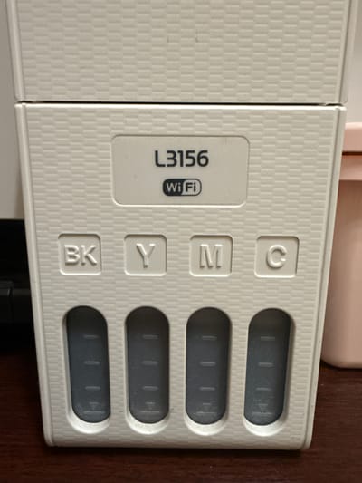 The image is a close-up, eye-level shot of the front panel of a white EPSON L3156 printer. The printer has a textured surface with a repeating grid pattern. Centered on the upper part of the panel is a white label with the text "L3156" in black, and below it, a black Wi-Fi symbol. To the left of the Wi-Fi symbol, the word "Wi" is in black, and to the right, "Fi" is in black.
Below the label are four recessed buttons, each with a single letter embossed in white. From left to right, these are "BK", "Y", "M", and "C".
Beneath these buttons are four oval-shaped ink tanks, also recessed. The tanks are filled with dark liquid, likely ink, and each has three horizontal lines and a downward-pointing arrow embossed on its side, indicating fill levels. The tanks appear to be partially filled, with the ink level below the top line in all visible tanks.
The printer is placed on a dark wooden surface, possibly a desk. To the right of the printer, a portion of a light pink container is visible. The background is out of focus, but appears to be a wall or another object. The lighting is neutral. The image does not contain any specific information about its location in Panevėžys, Lithuania.