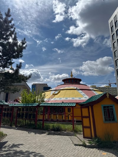 The image captures a section of the Gandantegchinlen Monastery in Ulaanbaatar, Mongolia. The foreground shows a brick pathway running along the right side. To the left of the path is a row of prayer wheels under a red and green roofed structure. Adjacent to this structure is the exterior of a yellow and red traditional building with intricate architectural details. In the background, modern buildings and a construction crane are visible against a bright blue sky with scattered clouds. A few trees are present in the lower-left corner.