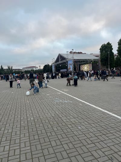 The image depicts an outdoor public event taking place in Kaspiysk, Russia, under an overcast sky during daytime. A large, temporary stage structure is central to the scene, featuring two prominent screens. The screen on the left displays an image of a person accompanied by the Russian text "СЧАСТЛИВОЕ ДЕТСТВО," which translates to "HAPPY CHILDHOOD," suggesting a thematic focus for the event. The other screen shows a vibrant, intricate patterned design.

Numerous people are gathered in front of the stage, forming lines and groups, seemingly watching a performance or presentation. In the foreground and midground, several children are present; one child is actively playing with a large white balloon on the paved ground, while another smaller child is also visible. A person on roller skates can be seen in the midground among the crowd.

The ground is paved with grey interlocking bricks, marked by a clear white line that extends into the distance, potentially indicating a track or designated area. Trees and distant buildings provide a backdrop to this urban outdoor setting. The overall atmosphere appears calm and engaged, with people gathered to observe the stage activities.