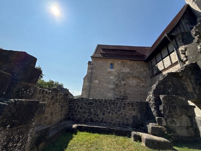 The image shows the exterior of the Walkenried Abbey in Walkenried, Germany, on a clear, sunny day. The foreground features rough, weathered stone structures, possibly remnants of older fortifications or foundations, covered in moss and lichen. These structures are low to the ground, with grassy areas interspersed between them.

In the midground, a well-preserved stone wall made of dark, irregular stones separates the foreground from the abbey building. Behind this wall stands the abbey itself, constructed from light-colored, roughly hewn stones. The abbey features a red tile roof with multiple layers of dormer windows or vents. A small, dark blue-paned window is visible on the stone facade. To the right of the main stone building, another section of the abbey is visible, with a dark wooden facade above the stone, suggesting a timber-framed structure or addition.

The sky is a bright, cloudless blue, with the sun prominently visible in the upper left quadrant, casting a strong glare and lens flare. The sunlight illuminates the scene, creating distinct shadows. In the background, a few trees and the rooftops of other buildings are visible, suggesting the abbey is situated within or near a village or town. No people are present in the image. The overall impression is one of historical architecture set against a bright, natural backdrop.