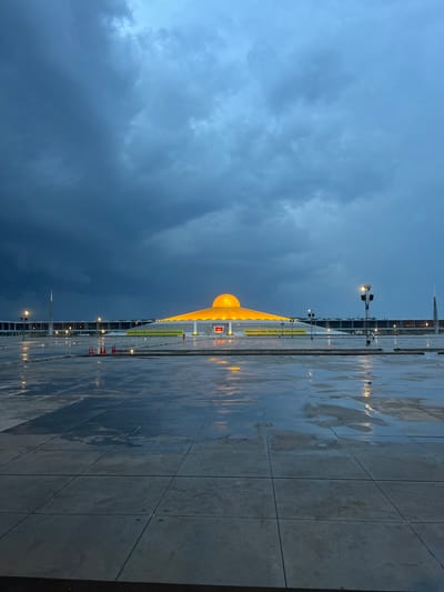 The image depicts the Dhammakaya Cetiya, also known as The Cetiya of One Million Lord Buddhas, located in Tha Khlong Town Municipality, Thailand. The structure is a large, golden dome illuminated from within, set against a dramatic sky filled with dark, turbulent clouds. The foreground consists of a vast, wet, tiled plaza, reflecting the ambient light from the structure and street lamps. Several traffic cones are visible on the plaza. In the background, a tall, slender spire and a long, low building with lights are visible. The overall atmosphere is moody and overcast, suggesting imminent or recent rainfall.