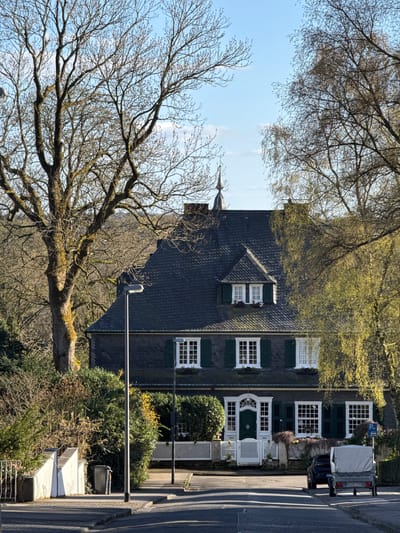 The image shows a three-story building in Remscheid, Germany. The building has a dark gray roof with white-framed windows and dark green shutters. A white picket fence surrounds the front of the house, and a white door with a decorative archway is visible. A vehicle with a trailer is parked in front of the building. A large tree with bare branches dominates the left side of the image. The sky is pale blue with some light cloud cover.