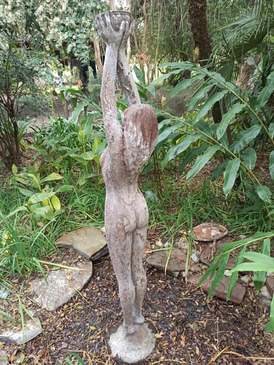 A weathered, nude female statue stands outdoors, centered in the frame. She is facing away from the viewer, with her arms raised above her head, holding what appears to be a bowl or vessel. The statue is textured and shows signs of age and outdoor exposure, with a reddish-brown patina. She stands on a round, rough base.

The statue is situated in a garden environment. The ground around her is covered with fallen leaves and twigs, interspersed with patches of grass and several flat stones of various sizes. Some of these stones are partially submerged in what appears to be standing water, suggesting recent rainfall or irrigation.

The background is lush with vegetation. To the left, a tree with delicate white blossoms is visible, and various green plants and foliage fill the middle ground. To the right, larger, broad-leafed plants dominate the scene, with palm-like fronds in the distance. The overall impression is a somewhat overgrown, naturalistic garden setting. The lighting suggests it is daytime, possibly overcast, with diffused light. The location is specified as Randburg, South Africa.