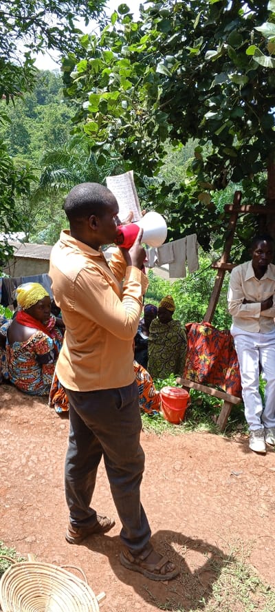 An African man in an orange shirt and gray pants speaks into a red and white megaphone, holding a notebook in his left hand. Several women in colorful patterned fabrics sit nearby. One man stands to the right, dressed in white. A red bucket, wooden structure, and hanging laundry appear in the background. The gathering takes place outdoors, with lush green trees overhead and a basket on the reddish-brown ground.