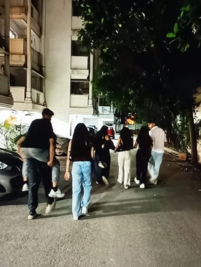 The image captures a nighttime scene in Aktash, Uzbekistan, depicting a group of seven individuals walking from left to right on a paved surface. On the far left, a male in a dark t-shirt and dark pants carries another male, dressed in a grey t-shirt and grey pants, piggyback style. To their right, a female wearing a dark top and light blue jeans walks. Further right, a cluster of three individuals, primarily seen from behind, proceeds; one is visible in a dark top and light-colored pants. The last individual on the right is a male wearing a light-colored shirt and light pants. A dark-colored vehicle is partially visible on the far left, next to the path. The background features a light-colored, multi-story building with balconies and columns on the left, and dense green foliage from trees or shrubs on the right. Artificial illumination casts shadows on the textured paved ground.
