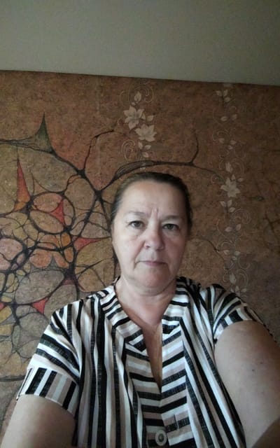 This is a medium-shot, front-facing photograph of an adult Caucasian woman. She has fair skin, brown eyes, and her hair is pulled back. She is wearing a horizontally striped shirt with black, white, and tan stripes. The background is a patterned wallpaper with abstract brown and beige organic shapes, possibly resembling veins or branches, with some lighter floral elements on the right side. The lighting suggests an indoor setting, possibly during the day. There is no visible text in the image. The woman's expression is neutral, and she is looking directly at the camera.