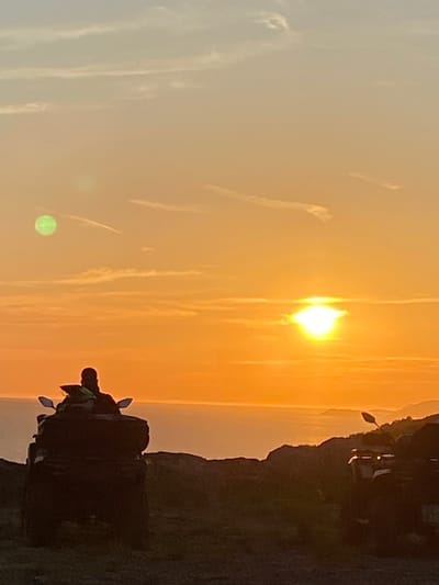 A man on a dark ATV faces away from the camera, looking towards the ocean at sunset. The sun is low on the horizon, casting a warm orange glow across the sky and water. The ATV is parked on a dirt path, with another ATV visible in the background. The scene appears to be set in a coastal area, possibly Montenegro, given the mention of Kovačko Polje. The sky is partly cloudy, with wispy clouds catching the sunlight. There is a hint of green light in the upper left quadrant of the image, possibly a lens flare.