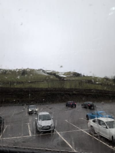 The image depicts an overcast, rainy day scene in Dolfor, United Kingdom, viewed through a rain-splashed window. The upper two-thirds of the frame show a dull grey sky with a faint, horizontal line of white dots across its center. Below the sky, a band of rolling hills with patches of green, brown, and white (possibly snow) extends across the mid-background, separated from the foreground by a dark strip of sparse trees or dense foliage.

The lower third of the image shows a wet asphalt parking lot with white painted lines marking individual spaces. Numerous water droplets are visible on the viewing surface, causing slight blurring. No individuals are present. Seven vehicles are visible in the parking area:
1.  A dark vehicle, partially visible on the far left foreground.
2.  A silver five-door hatchback (potentially a Ford Fiesta, license plate partially legible as "SG69 NLA") is parked facing right in the central foreground.
3.  A dark grey or black SUV with illuminated headlights is parked further back on the left, facing slightly right.
4.  A dark blue or black crossover SUV is parked in the mid-ground on the right, viewed from the rear.
5.  Another similar dark blue or black crossover SUV is parked further back on the far right, also viewed from the rear.
6.  A white sedan (possibly a BMW 3-series) is positioned in the foreground right, angled towards the left, partially within a parking bay.
7.  A bright blue sports car or performance hatchback is situated behind the white sedan, also angled towards the left.

The ground of the parking lot is visibly wet, reflecting light.