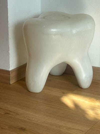 A white, tooth-shaped stool sits on a wooden floor in a corner of a room. The stool has a smooth, matte finish and three rounded legs that support a larger, crown-like upper section. The floor is made of light brown wooden planks with visible grain. A white wall with a wooden baseboard forms the background, meeting at a right angle. Sunlight casts a shadow across the floor to the right of the stool. The image appears to have been taken indoors, possibly in a home or office space. No people or actions are depicted. The location is not specified within the image itself.