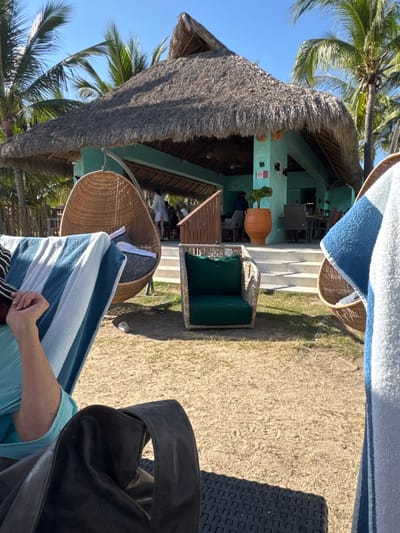 A tropical scene reveals a bar with a thatched roof, located on a grassy area with a view of palm trees under a bright blue sky. Wicker chairs with colorful cushions provide seating, along with a woven hanging chair draped with a striped towel. A section of dark patio furniture and a black bag occupy the foreground, obscuring the scene slightly.