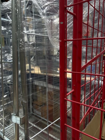 This media file presents a close-up, overhead view of a red metal shelf unit, likely in a commercial or storage setting. The shelf is covered with clear plastic shrink wrap, which obscures much of the detail of the items stored beneath. The overall impression is one of organized, yet potentially unboxed, inventory.
The red metal frame is composed of thick bars forming a grid-like structure. Some of these bars are horizontal, providing the shelving surface, while others are vertical supports. Diagonal bracing is also visible, adding to the structural integrity of the unit. The red paint appears to be matte.
Beneath the plastic wrap, the outlines of various objects can be discerned. These objects are also arranged on shelves, suggesting a multi-tiered storage system. Some of these items appear to be packaged in boxes or containers, while others may be more loosely arranged. The plastic wrap creates a somewhat hazy and reflective surface, distorting the view of the contents. The lighting appears to be artificial, typical of an indoor environment.
The presence of the red shelving unit, the clear plastic wrap, and the dense arrangement of items strongly suggest a retail stockroom, warehouse, or a similar storage facility. There are no people or specific environmental cues like weather or time of day visible in the image. The focus is entirely on the stored goods and the shelving system. There is some visible text on a white label peeking out from under the plastic wrap on the lowest visible shelf, with the letters "RI" clearly legible.
The location context provided is Holon, Israel, which could imply this is a retail or distribution center within that region. However, the image itself does not contain any specific visual markers that would definitively confirm this location.