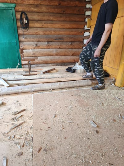In Islambaevo, Russia, a person stands in what appears to be an unfinished or construction area within a log cabin. The person is wearing a black t-shirt, camouflage pants, and patterned slip-on shoes. They are positioned near a yellow wooden pillar and a wall constructed of horizontal logs. The floor is covered with a thick layer of sawdust, wood shavings, and debris, along with several pieces of lumber and a rolled-up object wrapped in plastic sheeting. To the left, a green door is visible, and a small wooden chair with a metal base is also present. A coiled black cable hangs on the log wall near the door. The overall environment suggests ongoing work or renovation.