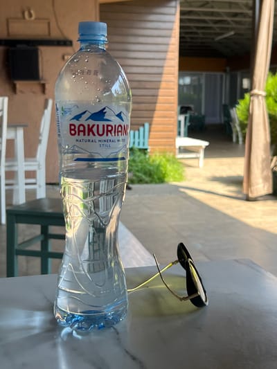 The image shows a clear plastic bottle of "BAKURANI NATURAL MINERAL WATER STILL" lying on a marble-like surface. The bottle is partially filled with water and has a blue cap. In the background, there is a wooden fence, some greenery, and outdoor furniture, suggesting an outdoor setting. There are also some structures that appear to be part of a building or covered area. To the right of the bottle, a pair of sunglasses with black frames and gold arms rests on the surface. The lighting suggests it is daytime, possibly late afternoon, with sunlight casting shadows. The scene appears to be calm and relaxed. The image is oriented vertically, with the bottle running horizontally across the frame.