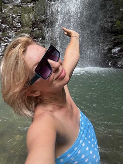 A woman with blonde hair and sunglasses poses in front of a waterfall. She is wearing a blue and white polka dot bikini top. The waterfall is cascading down a rocky cliff face into a pool of green water. The woman has her left arm raised, and her hand is reaching towards the waterfall. She is smiling and looking at the camera. The image is taken from a low angle, looking up at the woman and the waterfall.
