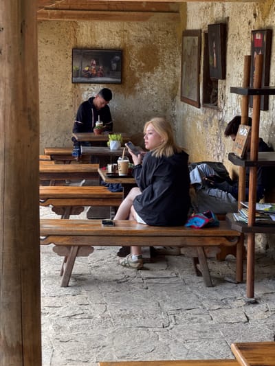 The image depicts an indoor scene in Lũng Cú Commune, Vietnam, with three people seated at adjacent wooden tables. A blonde woman wearing a black jacket and white shorts sits closest to the camera, using a mobile phone. A man in a black jacket is seated next to her, holding a drink. Further down the line of tables is another person, seated and engaged in their own activity. The tables are set with drinks and plants. The walls are a textured tan color, adorned with several framed pictures. The floor is made of grey stone. The shot is taken from a slightly elevated vantage point, partially framed by a wooden structure in the foreground.