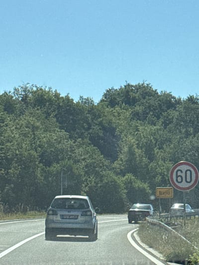 The image depicts a view from a vehicle traveling on a road, likely in Brajici, Montenegro, as suggested by a yellow road sign with the name "Brajici" on it. The scene is dominated by lush green trees forming a dense forest on either side of the road. In the foreground, a silver Volkswagen Golf is being driven on the right side of the road, with its rear visible. Further ahead on the road, another dark-colored vehicle is also visible. To the right of the road, a circular speed limit sign indicates "60". The sky is a clear, bright blue. The road itself is paved and has white lane markings. The overall impression is that of a scenic drive through a wooded area, with traffic present on the road.