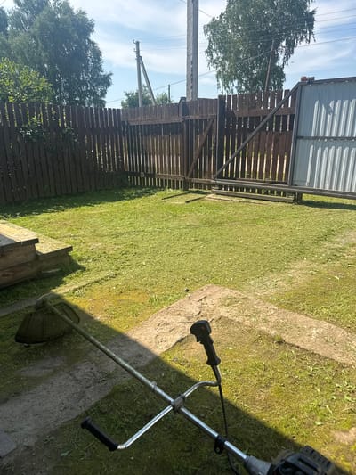 The image shows a backyard in Borodulino, Russia, with a recently mowed lawn. A string trimmer lies on the ground in the foreground, indicating that lawn maintenance has recently taken place or is about to. The yard is enclosed by a dark wooden fence. In the background, a corrugated metal gate and a utility pole are visible. The sky is bright blue with some clouds, and the sun is casting shadows across the lawn. There are no people or animals present in the image.