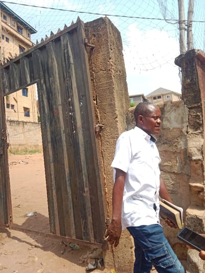 An adult male stands outdoors in Awada, Nigeria, facing right in profile. He wears a white short-sleeved collared shirt and blue jeans, holding a brown-covered book in his left hand. A second, unseen individual's hand is visible in the lower right foreground, holding a black smartphone directed towards the man. The man's right hand is positioned near this smartphone. Behind him, a tall, weathered, corrugated metal gate with spiked tops is partially open, revealing a dusty, unpaved area. To the left of the gate, a multi-story building with tan walls and dark windows is visible in the distance, partially obscured by a wire mesh fence overhead. To the right of the gate, a substantial, weathered concrete pillar forms part of an entrance, beyond which another deteriorating concrete wall is present. In the background to the far right, a residential building with a brown roof is visible. The ground is composed of reddish-brown dirt, with scattered debris near the gate's base. The sky above is bright and partly cloudy.
