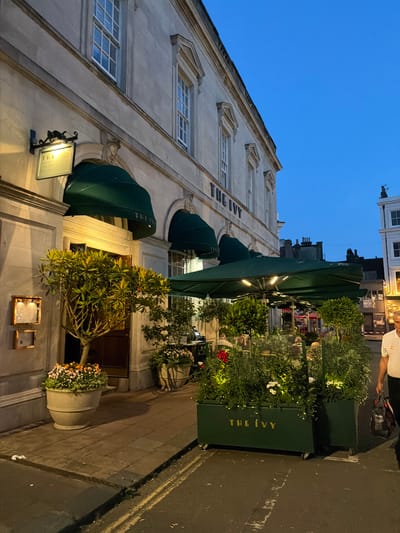 The image captures a nighttime scene on a street in Brighton, United Kingdom, outside a restaurant called "The Ivy." The building is illuminated by exterior lights, and the sky is a deep blue, indicating it's twilight or early evening. A sidewalk runs alongside the building, with planters filled with lush greenery and flowers lining the street.
The restaurant's facade features several arched awnings in a dark green color, suggesting an outdoor seating area. The building itself is constructed of light-colored stone with multiple windows. A sign with "THE IVY" written in gold letters is prominently displayed on one of the planters. Another sign, likely for the restaurant, hangs by a lamp near one of the entrances.
In the background, more of the street and buildings are visible, including a "TO LET" sign on another property. A few people are present, one walking by on the sidewalk, and others are presumably inside or associated with the restaurant, though their details are indistinct in the low light. The overall mood is calm and sophisticated, typical of an upscale dining establishment at dusk. The weather appears clear, allowing for the outdoor ambiance.