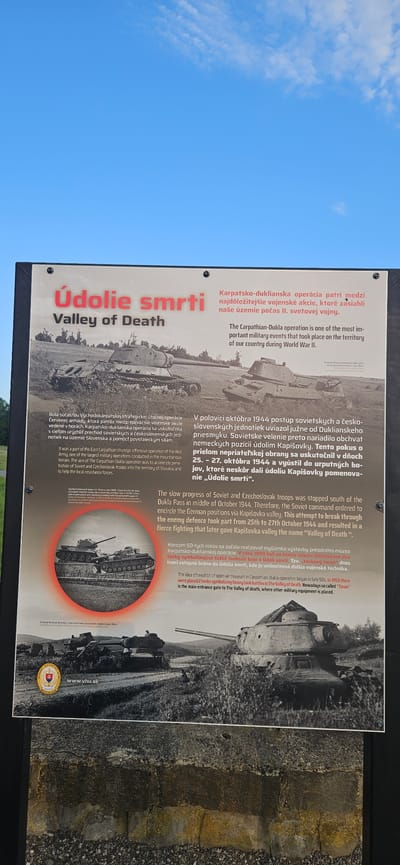 The image is a photograph of an informational plaque located outdoors, likely in the city of Svidník, Slovakia, given the textual content. The plaque is framed by a black border and displays historical information about the "Valley of Death" (Údolie smrti). The top section of the plaque features text in Slovak and English, with the Slovak text prominently displayed in red and white. Below the text are several black and white photographs depicting military scenes, primarily of tanks.

The main image on the plaque shows two Soviet tanks in a field, with a backdrop of rolling hills. Another smaller circular image, also in black and white, depicts a different tank scene. The text on the plaque details the Carpathian-Dukla operation during World War II, describing it as one of the most important military events on the territory of Slovakia. It mentions that Soviet and Czechoslovak troops were stopped by German defense in the Dukla Pass and that the fierce fighting gave the valley the name "Valley of Death." The plaque also mentions that an open-air museum was created in the Carpathian-Dukla operation area in 1959, with two tanks symbolizing heavy tank battles. The bottom portion of the image shows a concrete structure with gravel in front of it, which might be part of the museum or the display area. A Slovak emblem and the website "www.vhu.sk" are visible on the lower left side of the plaque. The sky is visible in the background above the plaque, with a few wispy clouds.