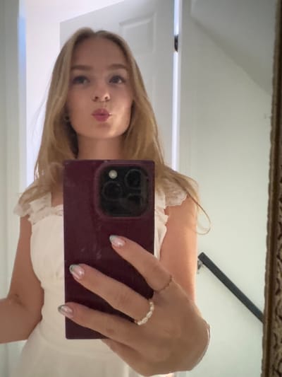 The image displays a mirror selfie of a female individual captured indoors. The subject, positioned centrally, has long, light blonde hair and is looking slightly upwards with puckered lips. She is wearing a light-colored, possibly white or off-white, dress featuring lace detailing on the shoulder straps. Her left hand, adorned with a thin band ring on the index finger and a multi-bead ring on the middle finger, holds a dark red or maroon mobile phone vertically, directed at the mirror. The fingernails on her left hand exhibit a light base polish with teal-colored tips. The reflected environment consists of light-colored walls, with a white door frame visible on the left side of the reflection. A dark handrail or banister is present on the lower right, indicating an interior space such as a hallway or staircase. A section of an ornate, gold-colored mirror frame is visible along the right edge of the image. The specific geographic location of Area B (Shawnigan Lake), Canada, is not discernible from the visual evidence provided.