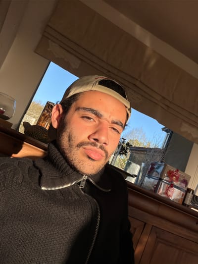 A selfie captured in a domestic interior, possibly in the city of Mauléon-d'Armagnac, France, based on the query. The subject is a young man with a beard, wearing a tan baseball cap and a black zip-up sweater over a turtleneck. He is positioned close to a window, with sunlight illuminating his face and the surrounding room. Outside the window, there are trees and the roof of a building is visible. On a shelf adjacent to the window, there are glass containers, possibly filled with items.