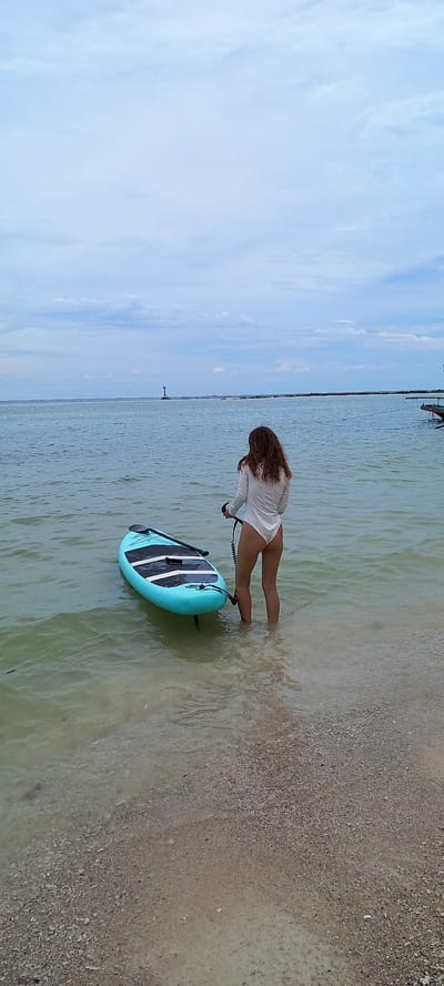 A young woman, waist-deep in shallow, slightly murky water, prepares to use a turquoise and black paddleboard. She is wearing a white long-sleeve shirt. The location appears to be a beach near Baan Chaloklum, Thailand, as evidenced by a distant navigational marker (likely a lighthouse) on the horizon. A strip of land is also visible in the distance. The sky is overcast with streaks of light blue. The beach is composed of light-colored sand and small pebbles.