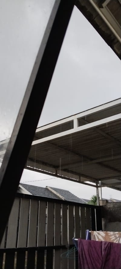 The image captures an outdoor scene, likely a residential area in Duman, Indonesia, on a cloudy and rainy day. A dark, triangular metal frame dominates the left side of the foreground, obscuring part of the view. Beyond this frame, a covered patio or carport with a corrugated metal roof is visible. The roof is supported by a white metal frame and shows signs of rain, with visible streaks of water.
Below the patio, a dark wooden fence with vertical slats separates the immediate area from what appears to be a tiled roof of a house. The tiles are dark grey. In the background, more roofs are discernible under the overcast sky. On the right side, clothes are hanging on a clothesline, including a purple garment and a patterned fabric, suggesting laundry is being dried or has been brought in due to the rain. A light green clothes hanger is also visible.
The weather is clearly inclement, with the sky being a uniform grey and rain falling, indicated by the streaks on the patio roof and potentially on the unseen window or surface through which the photo is taken. The overall mood is somewhat subdued due to the weather. There is no visible text in the image. The time of day is difficult to pinpoint precisely but the overcast conditions suggest it's likely daytime, possibly morning or afternoon. The scene evokes a feeling of a typical rainy day in a residential environment.