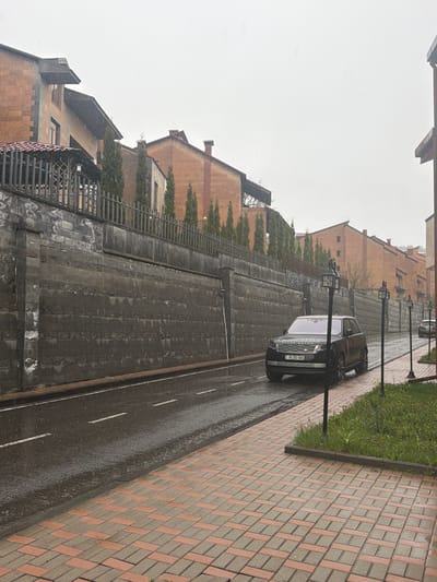 The image shows a wet street in Tsakhkadzor, Armenia. A black Range Rover is parked on the street next to a lamppost. The street has white lane markings. A brick walkway borders the street on the right, with a narrow strip of grass separating them. On the left, a stone retaining wall rises, topped with a row of small trees and iron railings. Buildings are visible behind the wall. The sky is overcast, and it appears to be raining or snowing lightly.