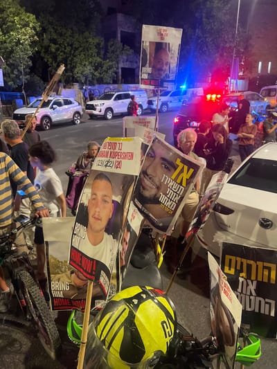 This night-time image depicts a public gathering or protest occurring on a street in West Jerusalem, Israel. The scene is illuminated by artificial light sources, including streetlights, vehicle headlights, and the flashing red and blue emergency lights of a police vehicle.

Approximately 15-20 individuals, primarily adults, are present in the foreground and mid-ground. They are engaged in a demonstration, holding numerous signs and posters. Many of these posters feature portraits of individuals, predominantly young men, with accompanying text. Prominently visible signs include:
*   A poster displaying a man identified as "Eitam Levi (33)" with the English text "BRING HIM HOME NOW!".
*   Another poster featuring a young man and text referencing "AVRAHAM KUPERSHTEIN (23) (22) (21)".
*   A third poster with a smiling young man and Hebrew text that includes "אל תשאירו אותי מאחור" (Do not leave me behind).
*   Other signs bear the English phrase "BRING HIM HOME NOW!" and Hebrew text on black backgrounds, such as "הורגים את החטופים" (Killing the hostages). Some also include hashtags like "#HOSTAGESFAMILIES".

People are standing and moving within the street area. One elderly woman is visible in the mid-ground, seated in a mobility device resembling a wheelchair.

Various vehicles are present:
*   In the background, multiple white civilian SUVs or pickup trucks are visible, one exhibiting an iridescent paint finish.
*   A marked police vehicle with active emergency lights is positioned in the mid-background.
*   A white sedan is partially visible on the right, with several protest signs leaning against its rear bumper.
*   In the immediate foreground, a motorcycle or scooter has a bright yellow and black helmet resting on its handlebars, with multiple signs attached or positioned nearby. A bicycle is partially visible on the far left, next to an individual.

The environment includes trees lining the street and a building facade visible in the background. The overall activity appears to be a vigil or protest advocating for the return of individuals referenced on the posters.