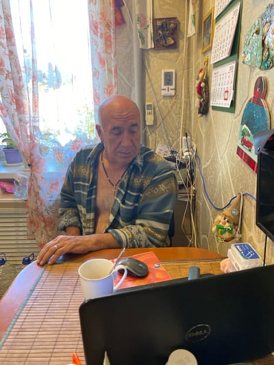 An elderly bald Caucasian man with his shirt unbuttoned is sitting at a table in front of a laptop in a room that appears to be a home office or living space. The room has stone-patterned walls, with several calendars and decorative items hanging on them. A window with floral curtains is visible to the left, with sunlight streaming through. On the table, there is a white mug with a spoon, a computer mouse, and a partially visible laptop. The man is wearing a necklace with a cross pendant. His left hand is resting on the table, and he appears to be looking down. The overall impression is of a casual, domestic setting. The image does not contain any elements that would allow for an identification of the location as Yokohama, Japan.