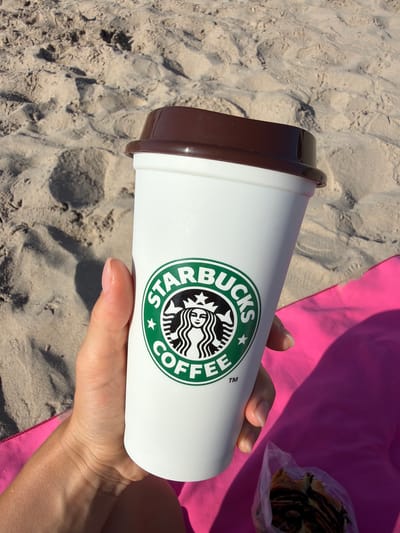 A person holds a white Starbucks coffee cup with a brown lid on a sandy beach. The Starbucks logo, featuring a siren, is visible on the cup. A bright pink towel is partially visible beneath the cup, and a plastic bag containing a pastry is in the lower right corner of the image. The background consists of rippled sand, indicating a beach environment. The image was taken in Finestrat, Spain.