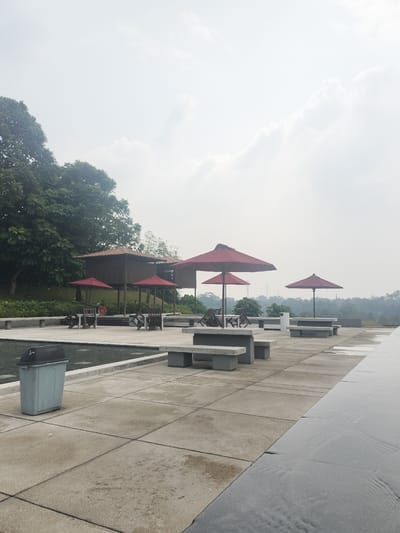 This is a photograph of an outdoor seating area with a pool or water feature in the foreground, located in Gunung Putri, Indonesia. The area features a tiled patio with several concrete benches and tables, some of which are covered by red umbrellas. In the background, there are two wooden structures with brown roofs, possibly gazebos or small buildings. A lush green forest is visible behind these structures, and in the distance, a hazy landscape with trees and possibly some industrial structures like power lines can be seen. The sky is overcast with diffused light. A light blue trash bin is positioned to the left of the frame, next to the water feature. One person in a red shirt is seated at a table under an umbrella.