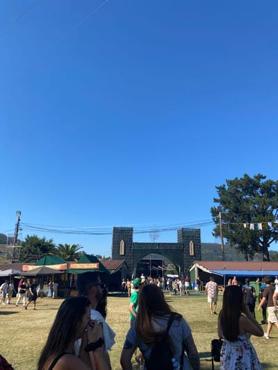 The image depicts a sunny outdoor event, likely a festival or fair, taking place in Llanera, Spain. In the foreground, several people are visible, some facing away from the camera, engaged in conversations or looking around. The middle ground features market stalls with green awnings, one with signage that appears to read "Tarta di Bella Carnal". Beyond the stalls, a large, dark green, castle-like entrance structure with arched openings is prominent. Attendees are milling about in front of this structure and the stalls. The background consists of trees and hills under a clear blue sky, with some power lines and a utility pole visible. The overall atmosphere suggests a daytime gathering with a festive or historical theme.