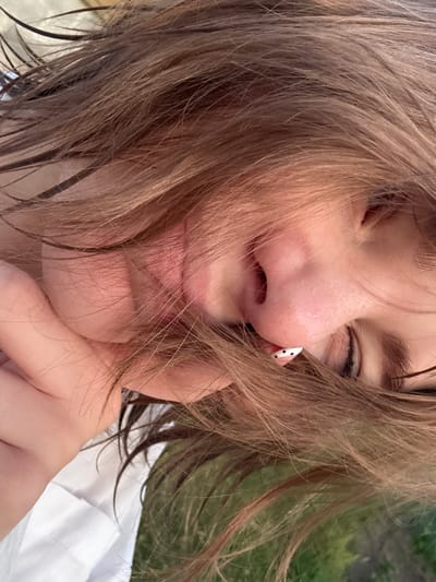 The media file is a close-up, slightly blurry selfie of a person's face. The subject's eyes are closed, and their hair, a light brown or sandy blonde color, is messy and falls over their face, obscuring parts of their features. The person is wearing white clothing, possibly a shirt or blouse, and has a hand resting on their chin. A small, black and white spotted earring is visible on their nose.
The setting appears to be outdoors, with a green, slightly blurred background suggesting grass or foliage. The lighting implies it could be daytime, possibly late afternoon or early evening given the soft light. The overall mood is relaxed and candid, with the closed eyes and hand gesture suggesting a moment of peace or perhaps playfulness. There is no visible text in the image. The location context provided is Ivatsevichy, Belarus.