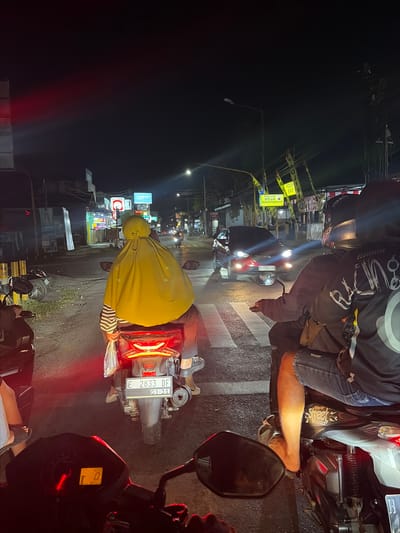 The image captures a nighttime scene on a street in Boja, Indonesia, characterized by a traffic intersection with visible streetlights, storefronts, and signage. In the foreground, the perspective is from the back of a motorcycle, showing its dashboard with a digital display indicating "0". To the immediate right, another motorcycle is partially visible, with a passenger wearing a black jacket and a helmet, their bare right leg and foot resting on the vehicle.

Dominating the center of the image is a third motorcycle, also viewed from the rear. This motorcycle has its brake lights illuminated, displaying the license plate "E 2833 DF G1-31". The rider is wearing a yellow headscarf that covers their entire head and shoulders, and a striped long-sleeved shirt. They are holding a plastic bag in their right hand. This motorcycle is positioned at a pedestrian crossing marked with white stripes on the asphalt.

In the background, a red car with its headlights on is approaching the intersection from the left. Several other motorcycles are visible further down the street. The environment includes illuminated storefronts with various advertisements, including "OPPO" logos. Yellow banners with the text "POLSEK BOJA" are prominently displayed on poles along the roadside, indicating the presence of a police station. The overall ambiance is that of a busy urban street at night.