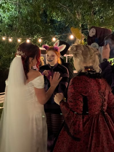 This evening photograph, taken in Nir Eliyahu, Israel, depicts a lively outdoor event, likely a themed party or wedding reception, illuminated by warm string lights. The background consists of dark green foliage.
In the center of the frame, a person dressed as the Cheshire Cat from Alice in Wonderland is prominently featured. They wear a purple and black striped top, purple cat ears, and have their face painted with whiskers and a wide, mischievous grin. They are looking towards a person in the foreground who has vibrant red hair and is wearing a white dress, possibly a bridal gown, along with a sparkling hair accessory.
Behind the Cheshire Cat, another individual is visible in a dark red dress with an ornate, possibly Victorian-era, pattern. Further back and to the left, parts of other costumed individuals can be seen; one appears to be wearing a light-colored wig and holding a small clock face, suggesting the character of the White Rabbit. The overall mood is festive and whimsical, characterized by the elaborate costumes and the soft glow of the artificial lighting. No text is visible in the image.