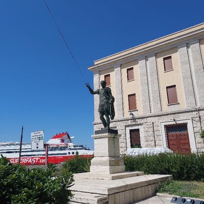 The image presents a daytime scene in Ancona, Italy, featuring the Statue of Trajan situated near the city's port. In the foreground, a bronze-colored statue of the Roman Emperor Trajan, depicted in full military attire with his right arm raised and left arm holding drapery, stands on a multi-tiered, light-colored stone pedestal. To the right of the statue, a classical-style building with a light-colored facade, distinguished by stone pilasters and multiple windows with closed reddish-brown louvered shutters, stands prominently. A large brown wooden door is visible on the building's ground level. Green bushes are present in the immediate foreground and along the base of the building. In the background, to the left of the statue, a large red and white passenger ferry, bearing the "SUPERFAST.COM" logo, is moored in the port. A white sign with black text and symbols is visible near the ferry. The sky is clear and blue, indicating bright, sunny conditions. A thin, dark line extends diagonally from the upper-left corner of the frame.
