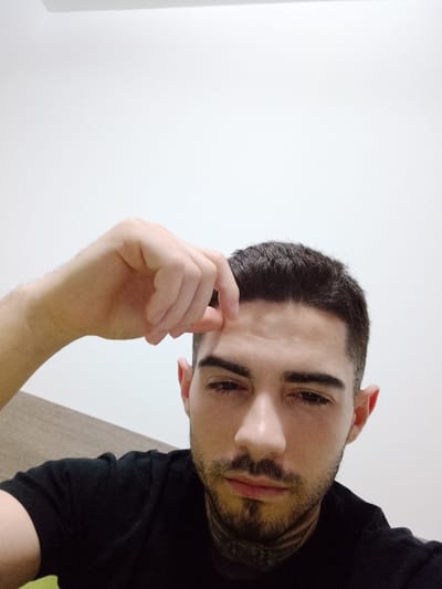 The image is a close-up selfie of a man indoors. He has short, dark hair, dark eyebrows, and a trimmed beard and mustache. He is wearing a black t-shirt. His right hand is raised, with his fingers touching his forehead. The background is a plain white wall. The location in the city of Fgura, Malta, is not ascertainable from the image.