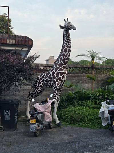 A large, full-body statue of a giraffe stands prominently in a courtyard, positioned next to a brick wall and lush greenery. The giraffe statue is depicted in a natural pose, with its head turned slightly to the right. In front of the giraffe, a parked scooter is visible, covered partially with a light pink cloth. To the left of the giraffe, a black trash bin with recycling symbols is placed on the paved ground. The background reveals a brick wall, dense foliage including a palm tree and smaller plants, and in the distance, trees and buildings under a cloudy sky. The scene appears to be an outdoor area in Xiaoshan District, China, possibly a residential or commercial space given the presence of the scooter and trash bin.