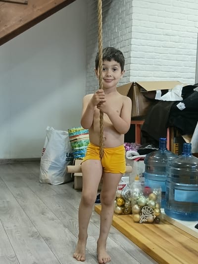 A young boy, likely under ten years old, is pictured suspended in mid-air, holding onto a thick rope. He is wearing bright yellow shorts. His body is positioned horizontally, with his arms gripping the rope and his legs extended. The boy has dark hair and a broad smile on his face, conveying a sense of joy or excitement.
The background is cluttered with various objects. To the left, a white brick wall is visible. To the right, stacked water cooler bottles and boxes of Christmas ornaments suggest a storage area or a room where decorations are kept. Several pieces of clothing are draped over furniture, including what appears to be a dark jacket and a white shirt. In the lower part of the image, a large white plastic bag is visible, along with a wooden structure. The floor is made of light-colored wooden planks.
The lighting appears to be artificial, suggesting the photo was taken indoors during the day or evening. There is no indication of the weather. The overall impression is of a playful moment captured in a somewhat untidy domestic or storage space. The text "UN" is partially visible on the white plastic bag. The location is Shcherbanka, Ukraine.