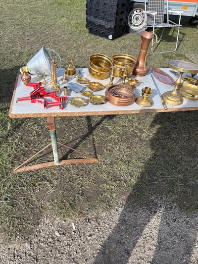 The image shows a selection of brass and copper items displayed on a table outdoors, likely at a market or sale in Chirk, United Kingdom. The objects include candlesticks, pots, a copper pitcher, and a metal bucket. Next to the table is a red metal contraption. The table is placed on a patch of mixed grass and dirt. To the right of the table, a tool with a rusty metal blade is stuck in the ground. The surrounding area is partly covered with gravel. In the background, a trailer is visible.