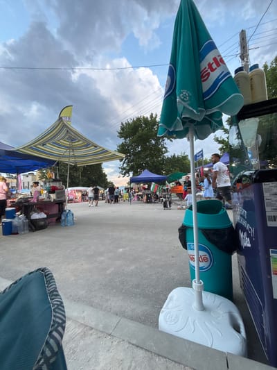 This image depicts an outdoor market or fair scene in Kostievo, Bulgaria, observed from a low vantage point, likely from a seated position. The setting appears to be an open-air pedestrian area with multiple vendor stalls under a partially cloudy sky. In the immediate foreground, a portion of a teal blue fabric chair with striped trim is visible on the bottom left. To the right, a large, closed teal blue sun umbrella with "Nestlé" branding in white and red lettering, along with a "MADE WITH RECYCLED PLASTIC" logo, is prominently displayed. The umbrella is secured by a white plastic base, resting on a light grey concrete or asphalt surface. Adjacent to the umbrella base is a teal green cylindrical bin also bearing the "Nestlé" logo, with a black trash bag visible at its opening. To the right of the bin, a dark blue commercial refrigerator or freezer is partially visible, displaying Cyrillic text "ОБИТ В НЕЖНОСТ! Milka" (Lived in tenderness! Milka) and an energy efficiency label. Two light-colored plastic bottles are placed on top of this refrigerator. In the midground to the left, a market stall features a distinctive striped canopy in shades of yellow, blue, green, and white. Above this canopy, a yellow banner-style flag is partially visible, showing the text "NCAKE" (possibly part of "PANCAKE"). Beneath the striped canopy, a woman in a light-colored shirt stands behind a counter, possibly engaged in food preparation or service, with various containers and a blender-like machine containing red liquid on the table. Several large plastic water bottles and blue coolers are arranged on the ground beneath the counter. A blue canopy from an adjacent stall is visible further to the left. The central and right midground shows a paved pathway extending into the distance, flanked by numerous market stalls. People are seen walking, standing, and interacting among the stalls. A man in a white t-shirt and dark shorts walks towards the right, followed by a young girl in a light blue dress. Near one of the stalls on the right, a signpost displays Cyrillic text "ПЛОВДИВ Plovdiv," indicating proximity or direction to the city of Plovdiv, and another sign below reads "БЪЛГАРСКА ЦАРЕВИЦА НА МАСА" (Bulgarian Corn on the Cob). A Bulgarian flag is visible at one of the stalls. Further down the pathway, a denser crowd of people is gathered around additional market setups. Trees line the background on both