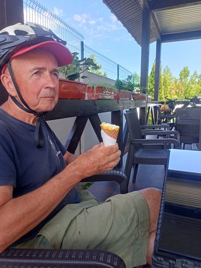 An elderly man wearing a cycling helmet and a dark blue t-shirt is sitting on a patio and eating an ice cream cone. He is holding the cone in his right hand. He is wearing green shorts. The man is looking to his left. The patio has a wooden roof and is furnished with wicker chairs and tables. Behind the man, there are potted plants on a wooden shelf, a metal fence, and trees. In the background, there are two bicycles. The scene appears to be outdoors on a sunny day. The location is the city of Shabo, Ukraine.