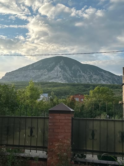 The image shows a natural landscape with a large mountain in the background. The mountain has a rugged, rocky surface. The foreground features a green area with trees, and a building, possibly a house, is visible amidst the foliage. A fence, made of metal and translucent material, is prominent in the right side of the image. The sky is partly cloudy, with some bright areas suggesting it might be daytime. A thin wire stretches across the frame from the top left. The overall scene appears to be a rural or semi-rural setting in a mountainous region. There is no visible text in the image. The orientation of the image suggests it may have been taken on its side.