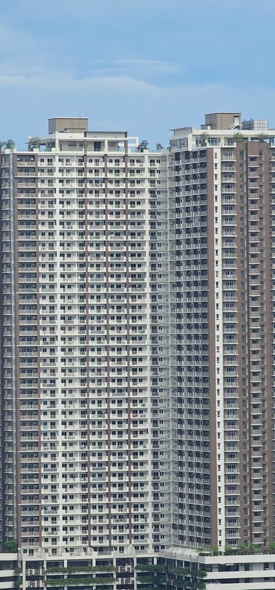 The image shows a close-up, full-frame view of a very tall, modern condominium building in Makati City, Philippines, against a pale blue sky with a few wispy clouds. The building's facade is primarily white with dark brown vertical structural elements and many balconies with white railings. The structure is composed of numerous identical floors, each featuring a repeating pattern of windows and balconies. Air conditioning units are visible mounted on many of the balconies. At the top of the building, there are rooftop amenities, including what appear to be white structures, possibly housing mechanical equipment, and some greenery. The lower section of the building, visible at the bottom of the frame, shows a different architectural style with more dark-colored elements and lush green landscaping. The overall impression is of a dense urban environment with high-rise residential architecture.