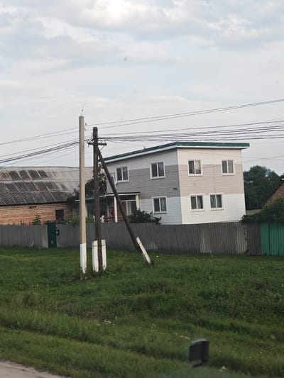 In the village of Chemodanovka, Russia, a two-story house with a light-colored facade and a green roof stands behind a grey metal fence. The house has multiple windows on both floors. To the left of the house, a corrugated metal barn with a brick base is visible, also behind the fence. A utility pole with wires runs across the foreground, leaning at an angle. The ground is covered in green grass. The sky is overcast with grey clouds.