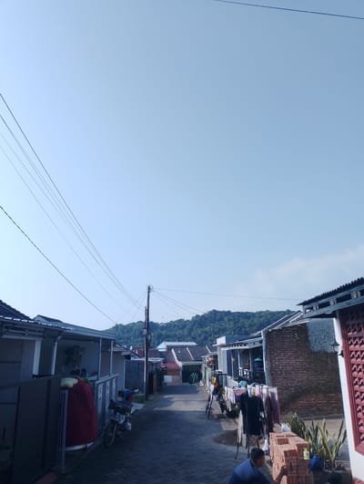 The image depicts a street scene in a residential area, possibly Kuningan, Indonesia, under a clear blue sky. The street is paved with interlocking bricks and lined with houses. Several houses have visible roofs, some with dark tiles and decorative eaves. One house on the left features a covered porch with columns and what appears to be a motorcycle parked nearby. Clothes are hanging on a rack in front of another house further down the street, suggesting daily life activities. There are also stacks of bricks visible in the foreground and near the houses, indicating ongoing construction or renovation. A person in a blue shirt is seated near the bricks, and another person in the background is attending to something near a motorcycle. Power lines crisscross the sky, supported by a utility pole. In the distance, a lush, tree-covered hill or mountain range forms the backdrop to the settlement. The overall atmosphere is one of a tranquil, rural or suburban Indonesian neighborhood.